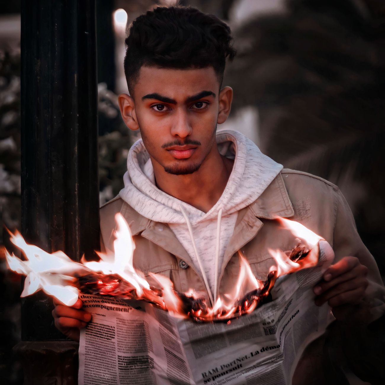 Portrait of a young man holding a burning newspaper outdoors, expressing intense emotion.