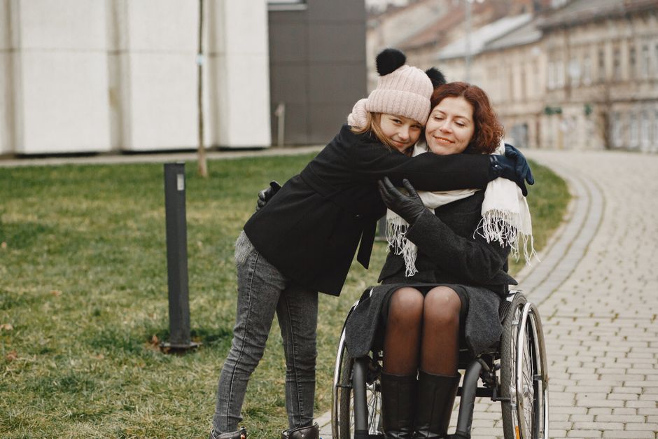 A young girl joyfully hugs a woman in a wheelchair outdoors, symbolizing love and care.