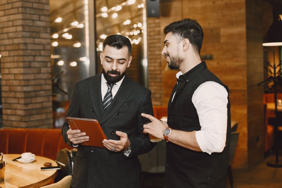Two businessmen engaging in a discussion while reviewing information on a tablet in an elegant lounge setting. Two businessmen engaging in a discussion while reviewing information on a tablet in an elegant lounge setting.