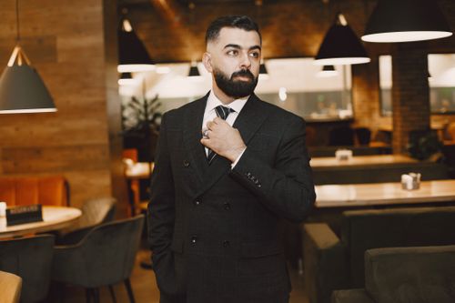 Free Stylish man in a black suit and tie standing confidently in a modern restaurant setting. Stock Photo