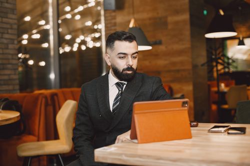 Free Man in business attire working on a tablet in a modern café with warm lighting. Stock Photo