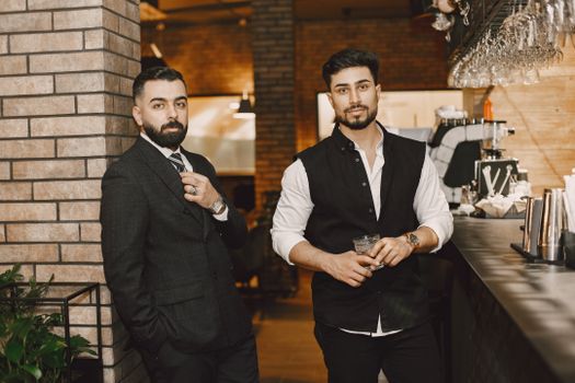 Two men in smart attire pose confidently by a trendy bar counter.