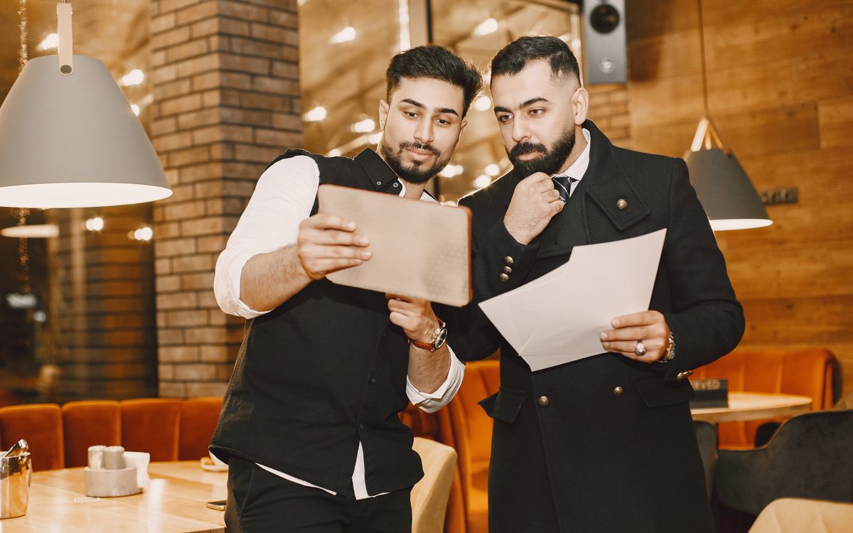 Two men in a restaurant review business documents and a tablet for planning purposes.
