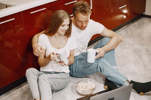Free A couple relaxing on the kitchen floor, enjoying drinks and watching a laptop. Stock Photo
