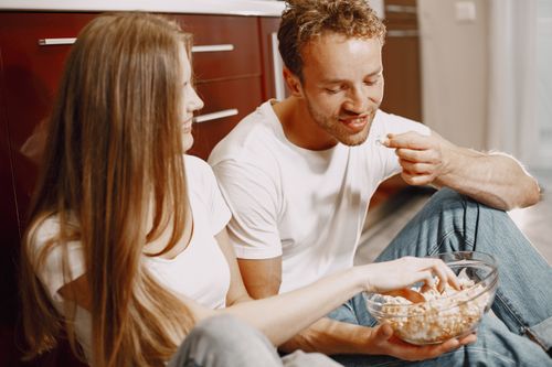 Free Happy couple sitting on the floor indoors, sharing a bowl of popcorn and smiling at each other. Stock Photo