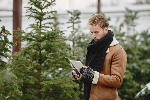 A man in a winter coat and scarf uses a tablet in a conifer tree nursery during the day.