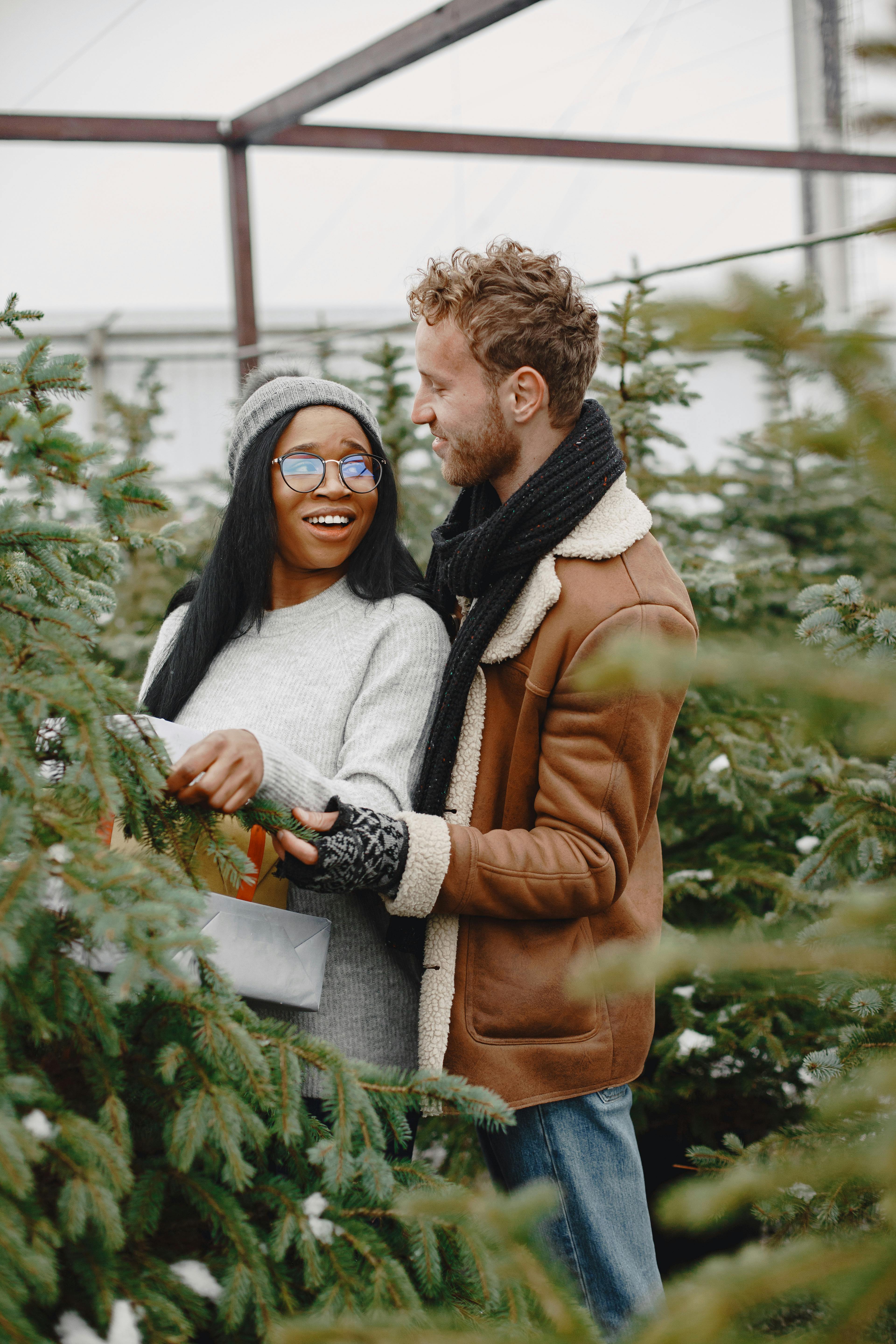 Joyful couple selecting a Christmas tree at a winter market, surrounded by festive greenery.