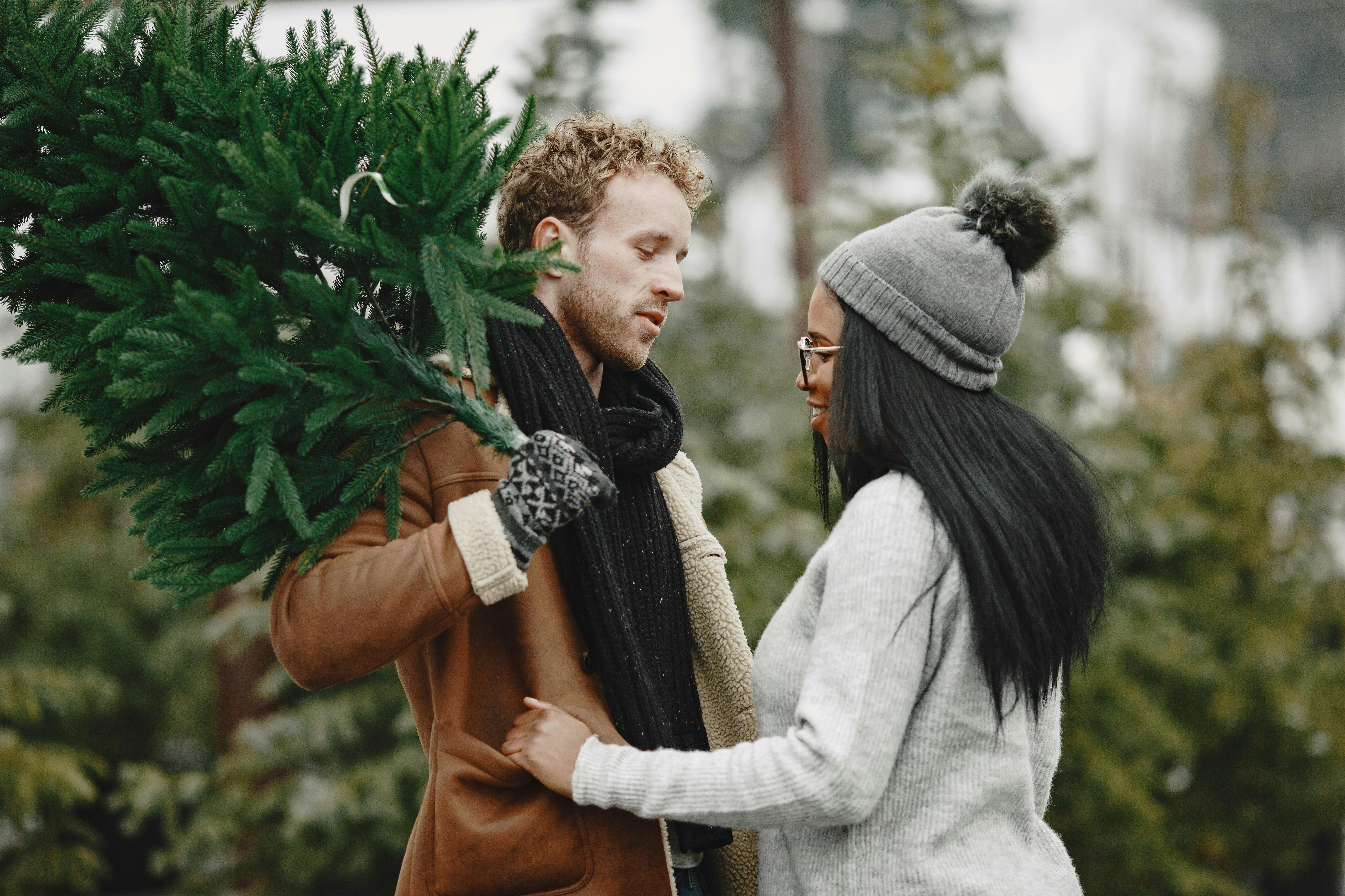 A couple enjoying a winter day, holding a Christmas tree amidst snowy conifer trees.