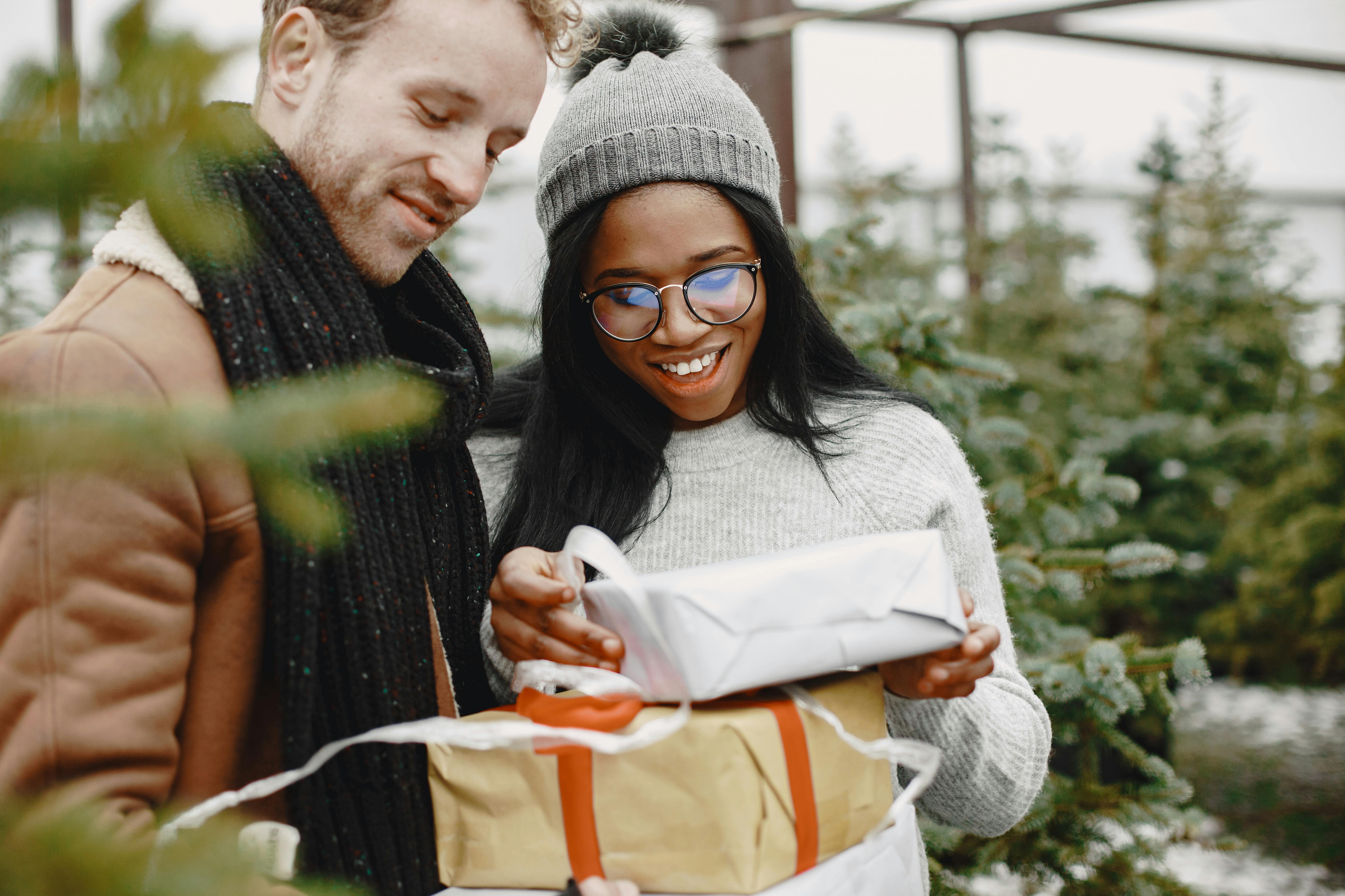 Joyful couple exchanging gifts surrounded by Christmas trees in an outdoor winter setting.