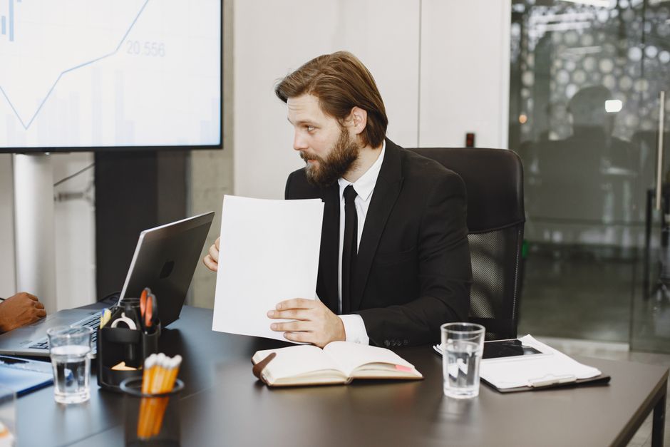 Um homem de negócios em um terno revisando documentos em uma mesa de escritório com laptops e blocos de notas.