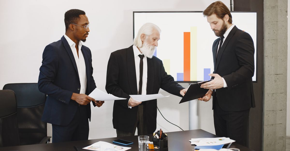 Three businessmen discussing documents in a conference setting with a presentation screen.