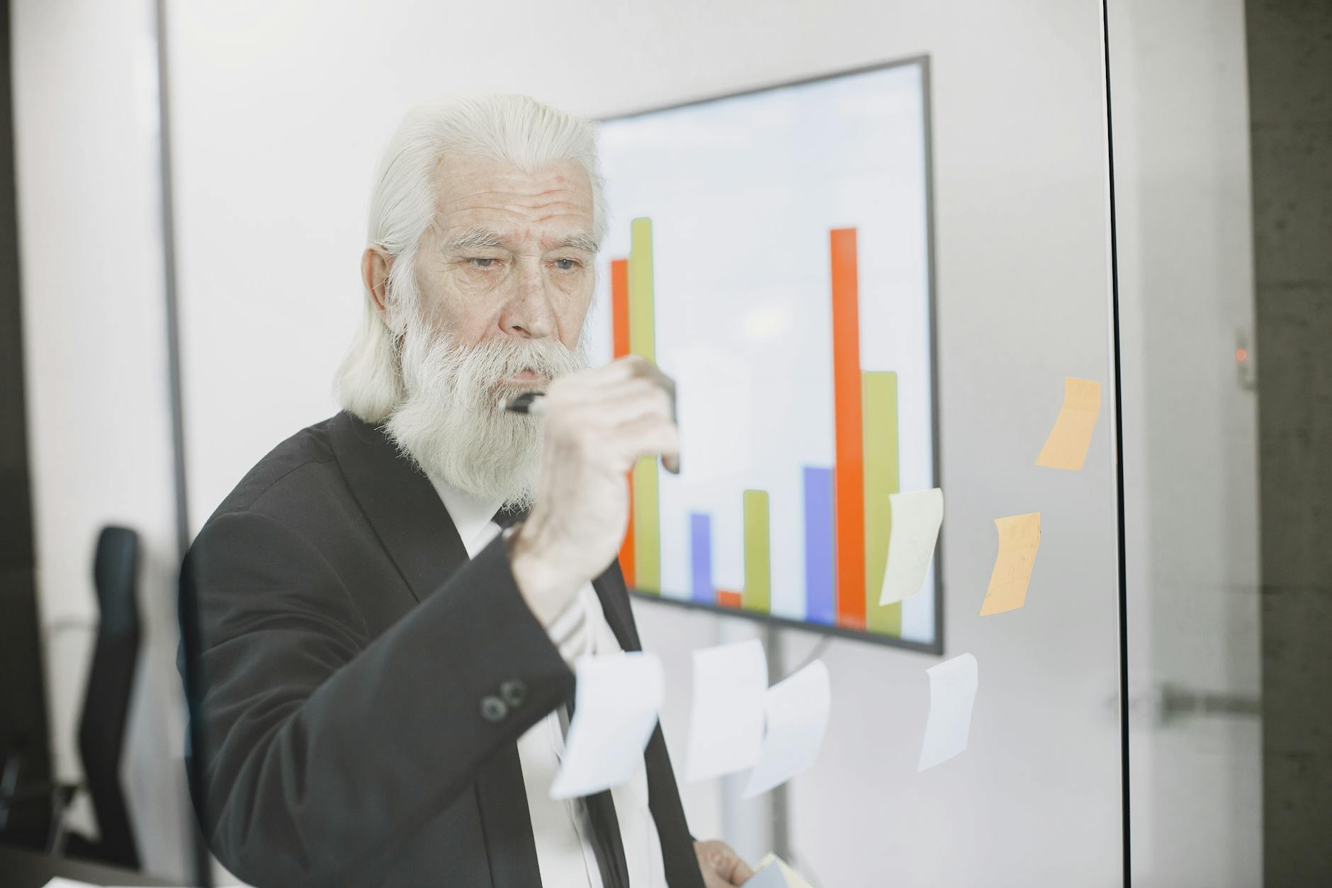 An elderly businessman in a suit writes on a transparent board with graphs in a modern office setting.