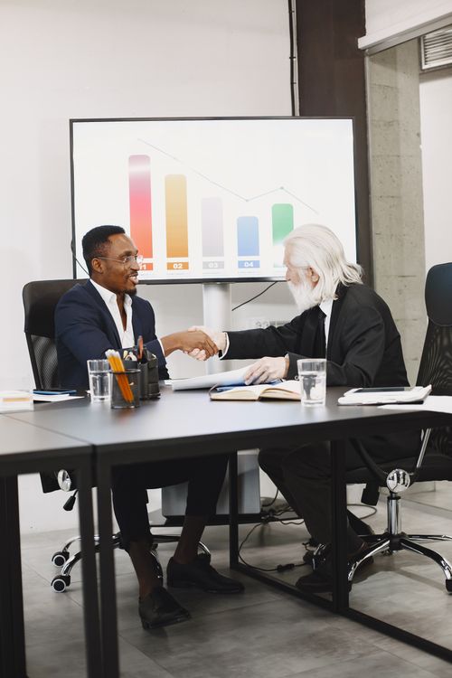 Free Two diverse businessmen in suits shaking hands over a desk in a modern office. Stock Photo