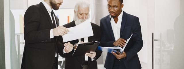 Three diverse businessmen in suits collaborate over documents in a modern office setting.