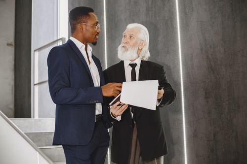 Free Two businessmen in suits discussing documents indoors on modern stairs. Stock Photo
