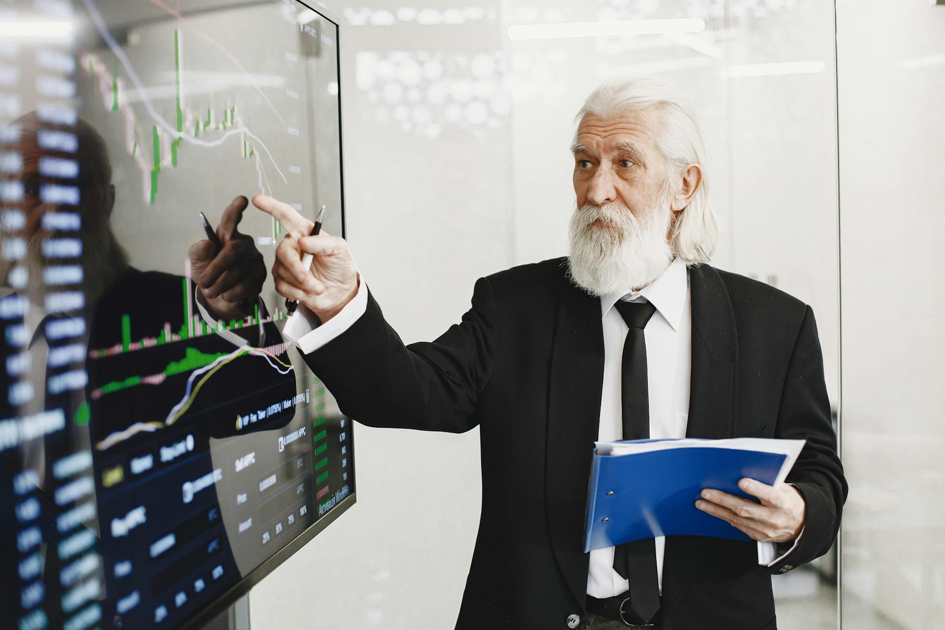 Elderly businessman pointing at stock market charts in an office setting, highlighting data analysis.