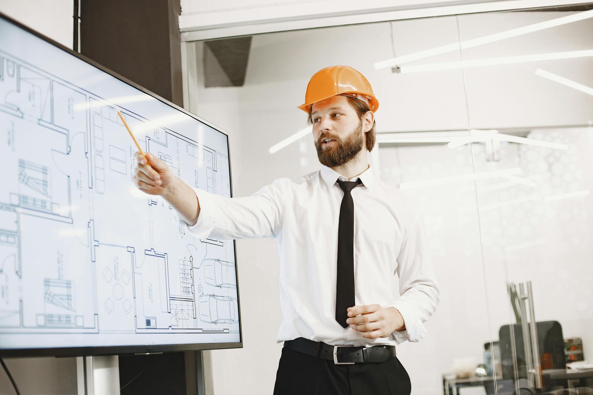 Architect with helmet presenting building blueprints on screen in modern office environment.