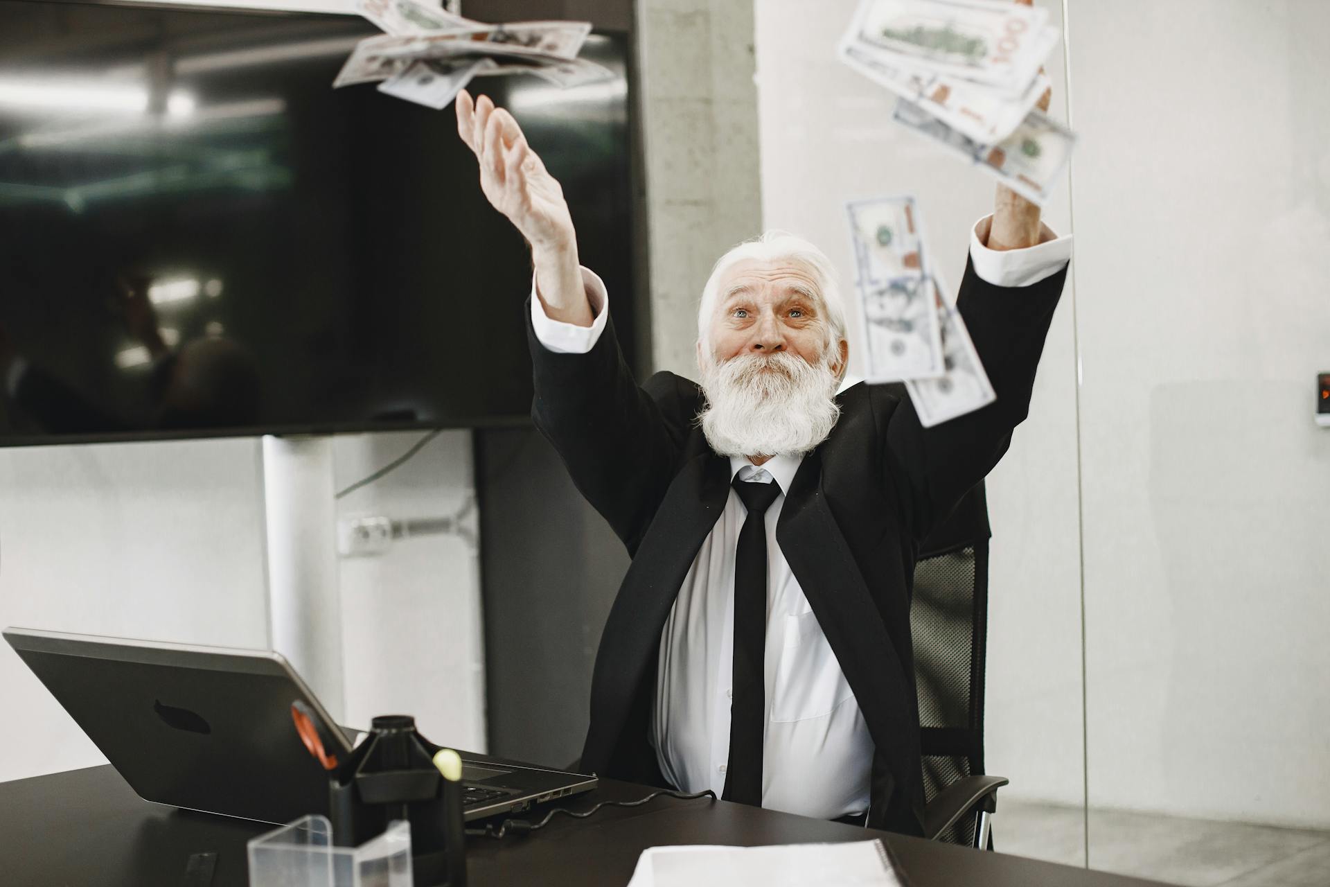 A joyful senior businessman in a suit throws money in an office setting, expressing financial triumph.