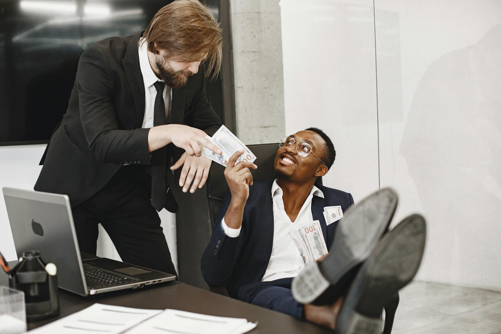 Two businessmen in suits exchanging money with smiles in a modern office environment.