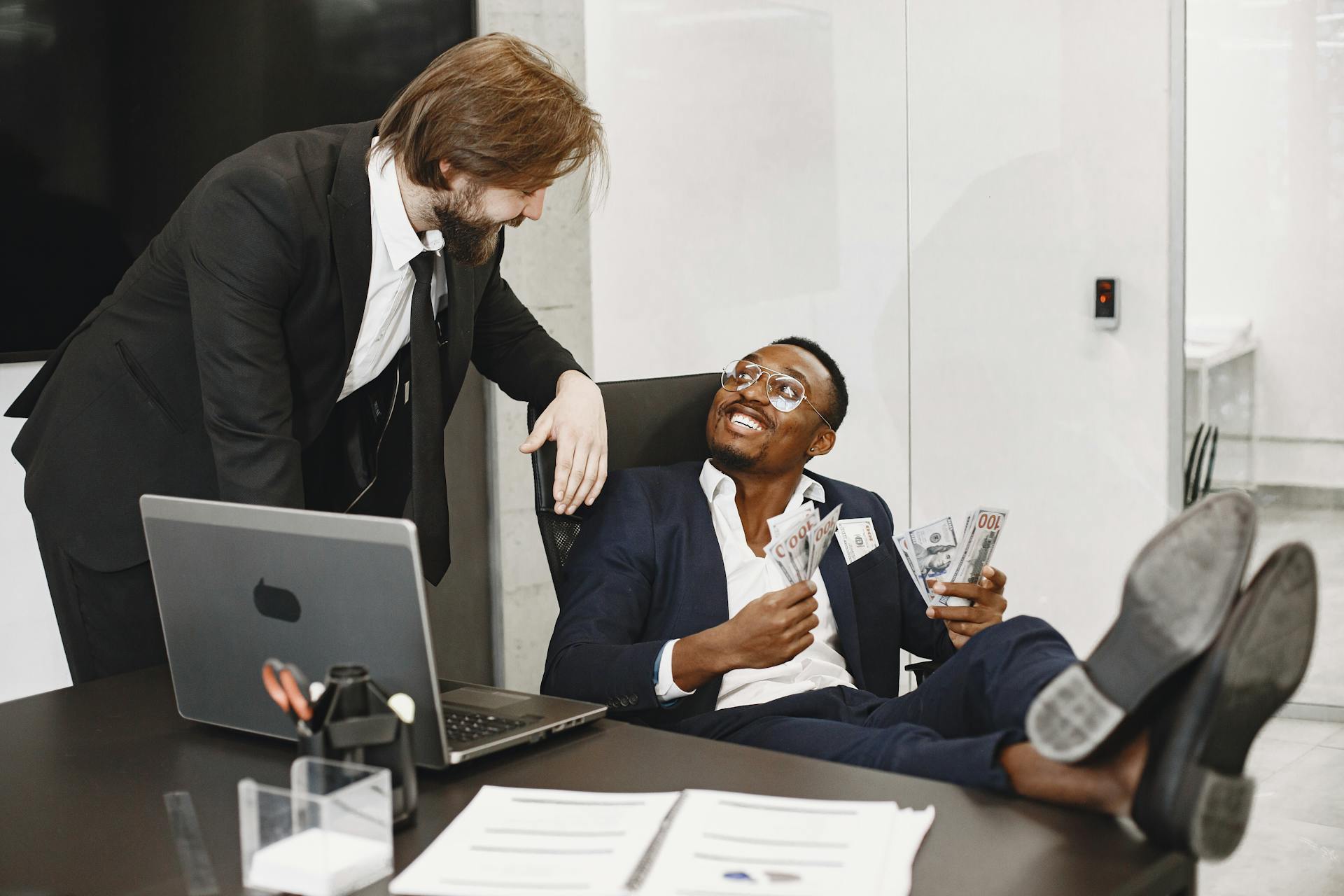 Two businessmen in suits celebrating a financial windfall at an office desk with cash.