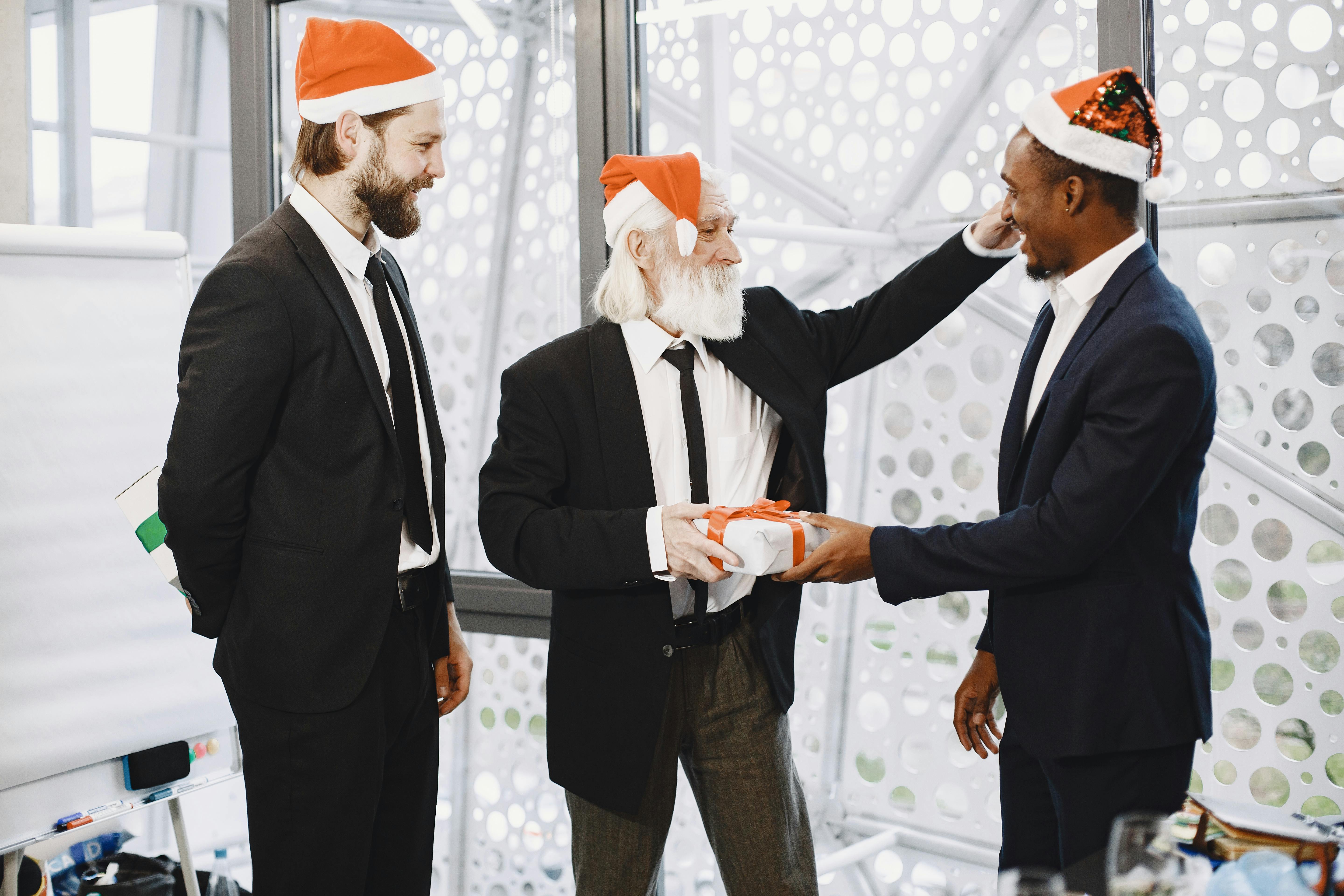 Three colleagues in suits exchange Christmas gifts in a festive office setting.