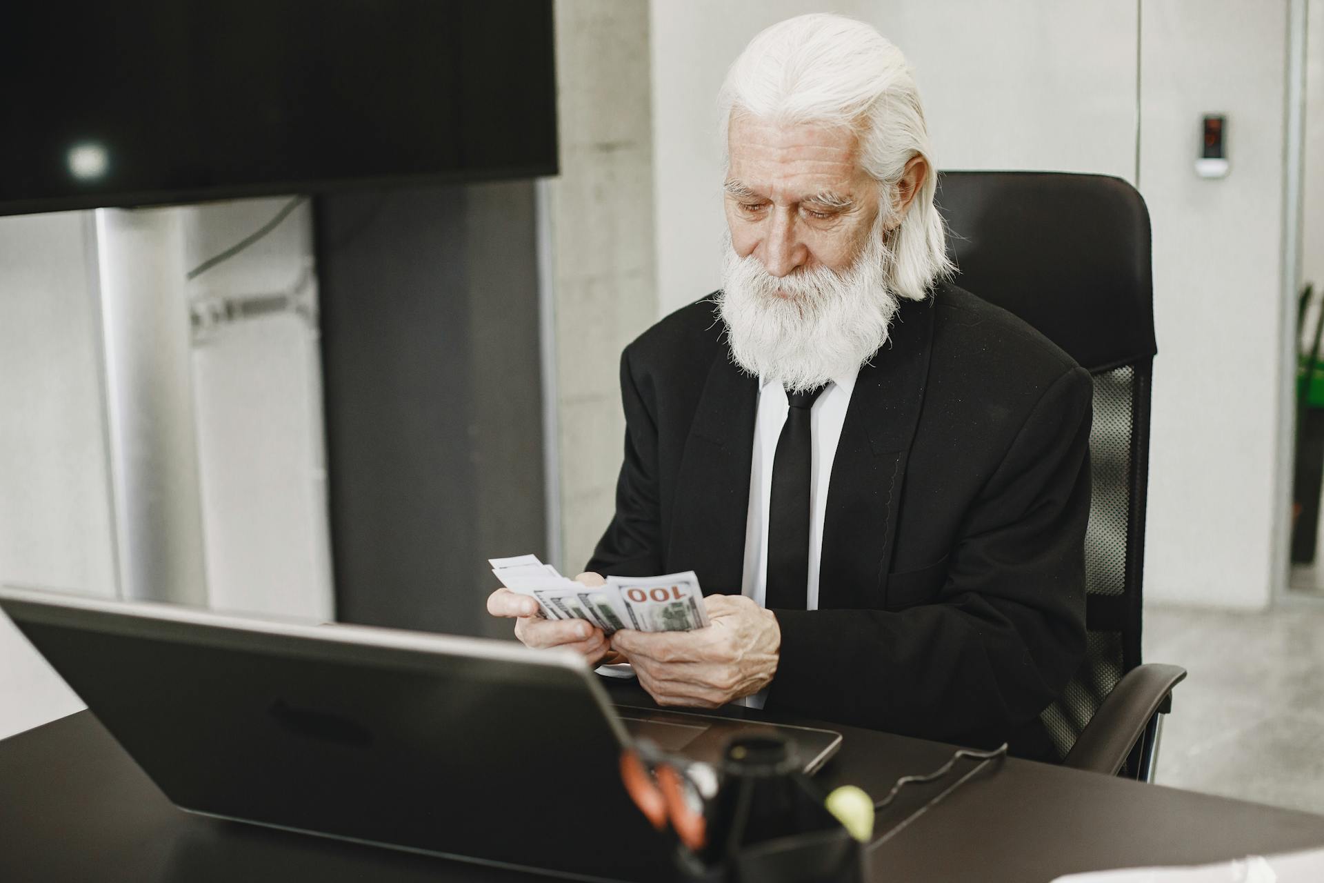 Senior businessman in suit counting money at office desk with a laptop. Elegance and financial focus in modern workspace.