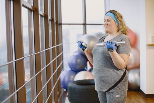 Woman in gym with dumbbells by a window, highlighting fitness and lifestyle.