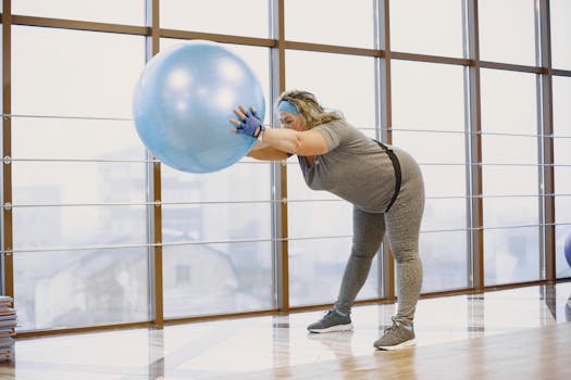 A plus size woman stretches using a fitness ball in a bright gym, promoting wellbeing and body positivity.