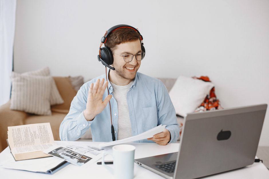 Smiling man in headphones holding paper and waving during a video call at home. Smiling man in headphones holding paper and waving during a video call at home.