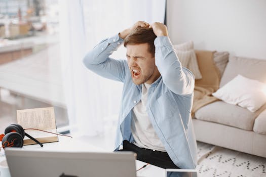 An upset man with mouth open, pulling hair at a desk with a laptop in a home setting.