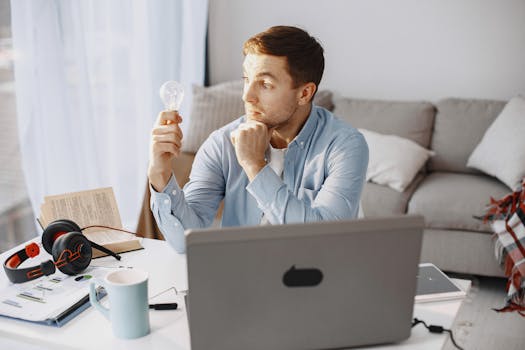 Young man at home desk holding a lightbulb, symbolizing a moment of inspiration during remote work.