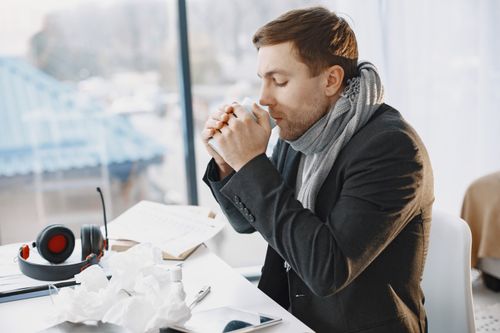 Free A man in a black suit and scarf drinks coffee in a bright office, suggesting he's unwell. Stock Photo