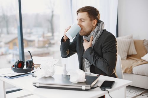 Free Man feeling unwell sitting at desk drinking from a mug with scattered tissues. Stock Photo
