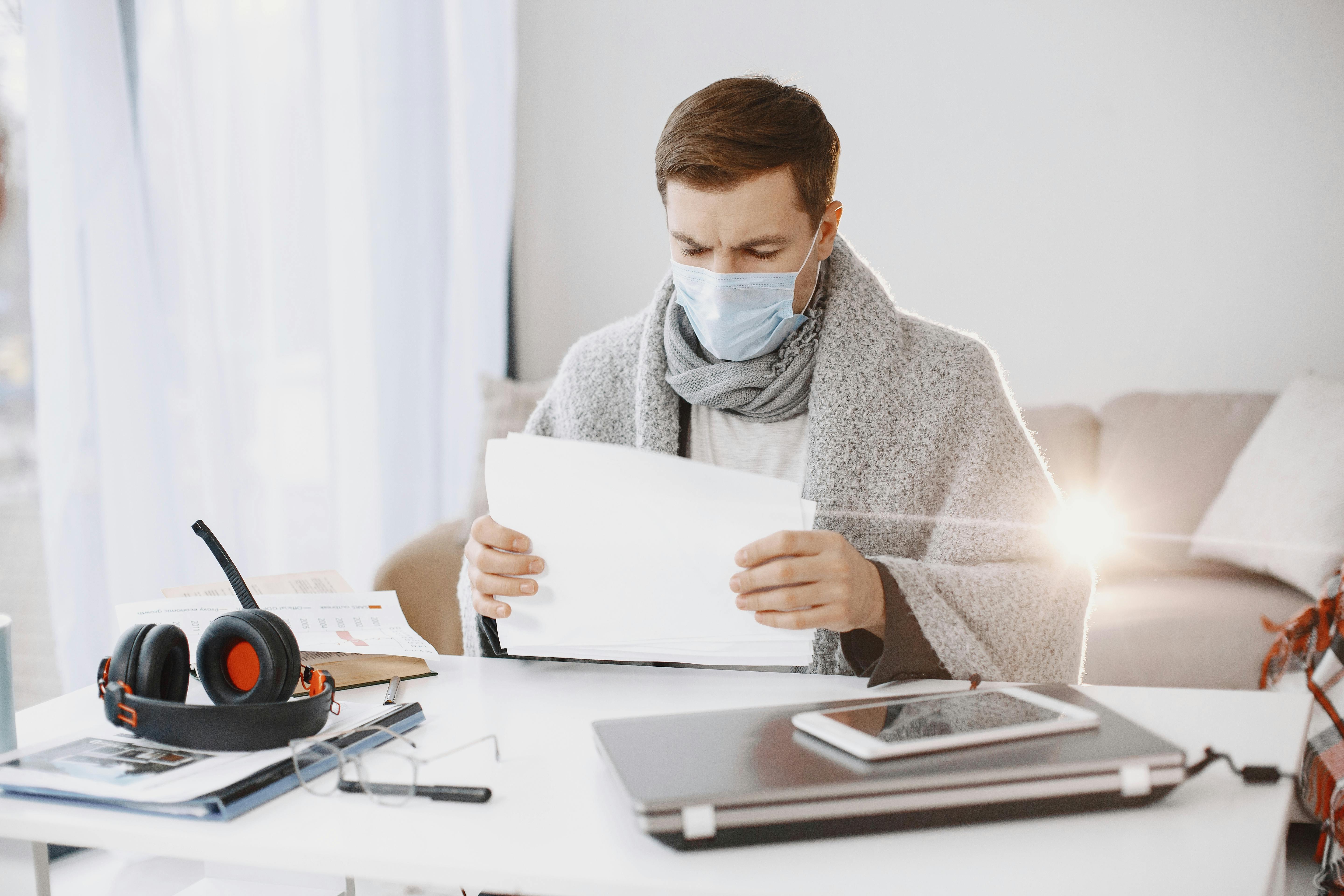 A man wearing a protective mask and scarf works from home, surrounded by office supplies on a bright