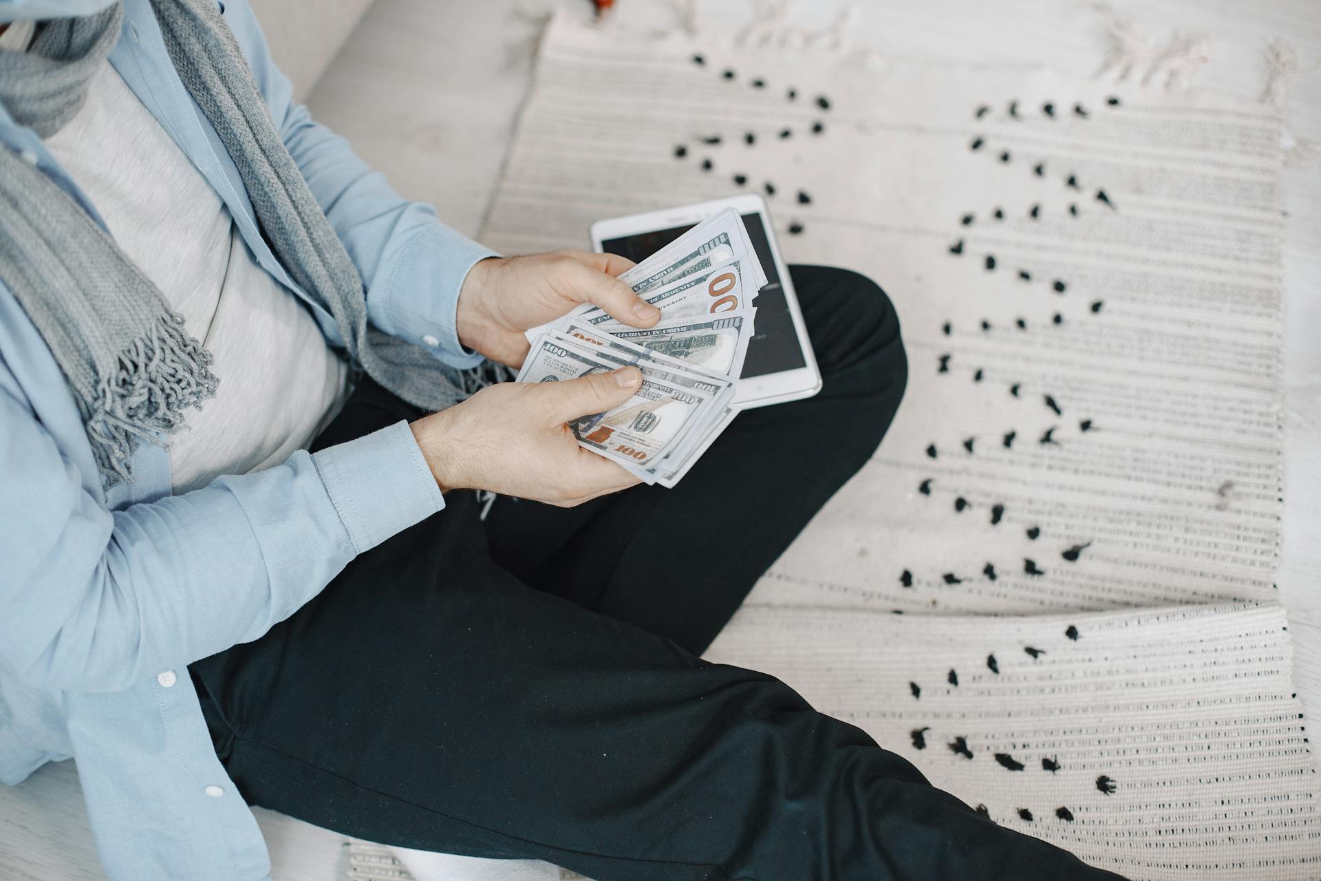 Close-up of adult counting cash with a tablet indoors, symbolizing finance and technology.