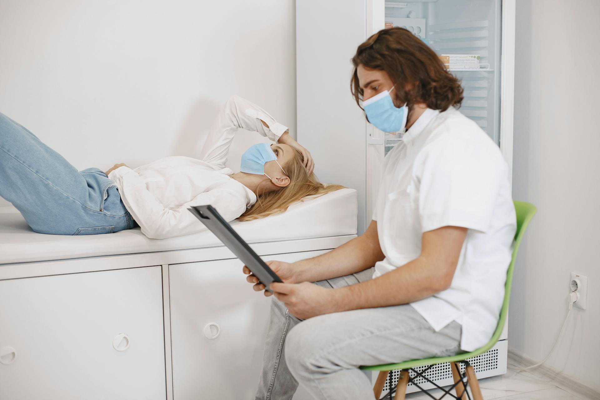 Doctor consulting patient wearing face masks in a medical examination room, discussing healthcare plan.