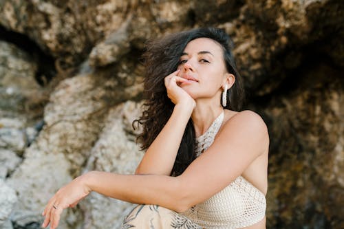 Low angle of young dreamy female dressed in boho style sitting in mountain terrain in sunny summer day