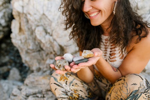 Smiling female with long curly hair dressed in boho style sitting in rocky terrain with stones pebbles and minerals in hands