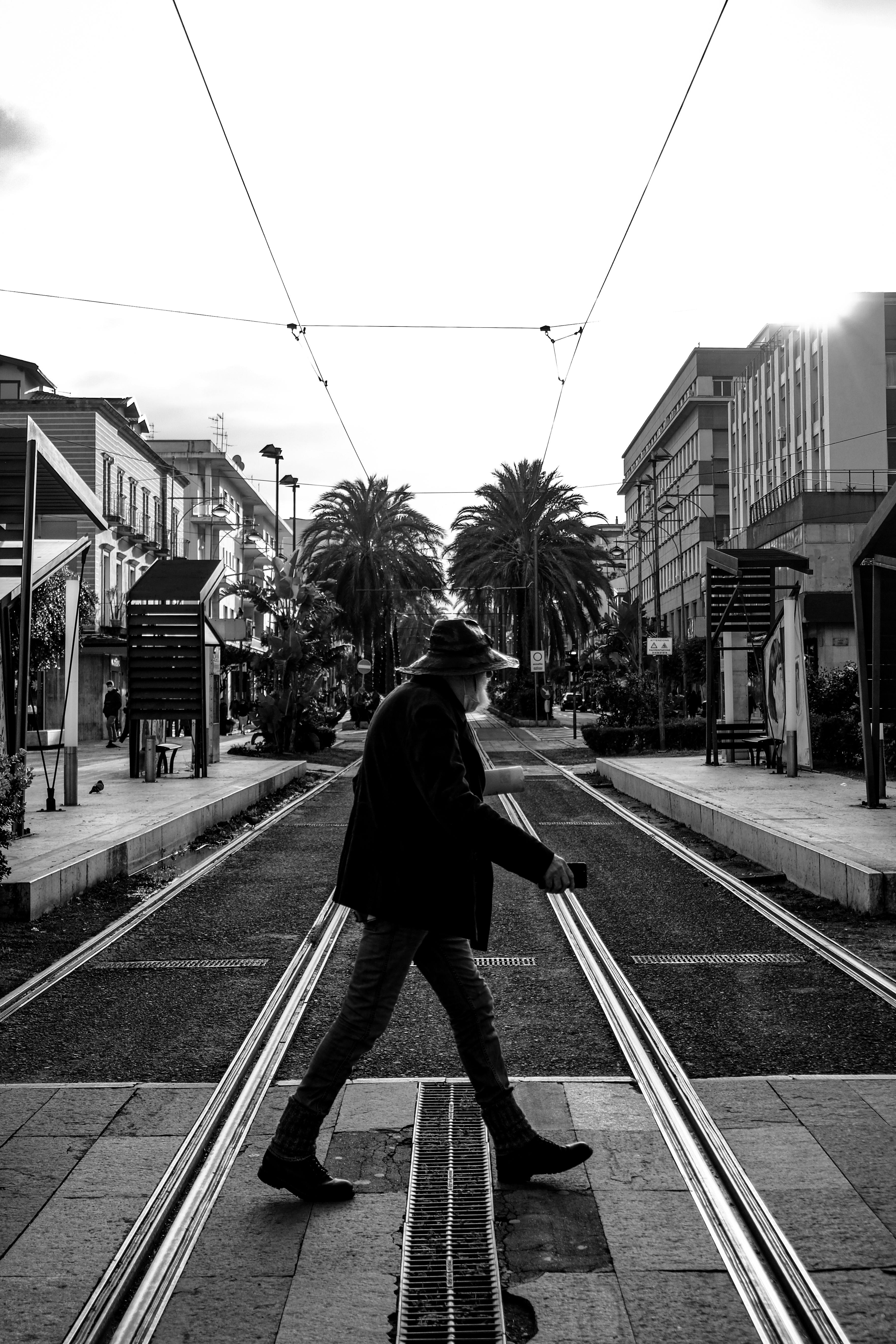 Black and white photo of a man crossing street with tramlines, tall buildings, and palm trees.