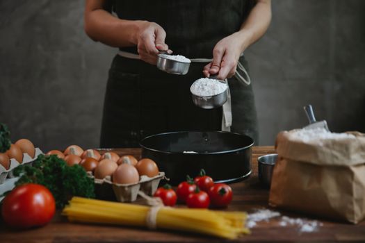 Crop faceless person in black apron standing near counter and adding flour in bowl while making homemade pasta with eggs and spaghetti near tomatoes and herbs
