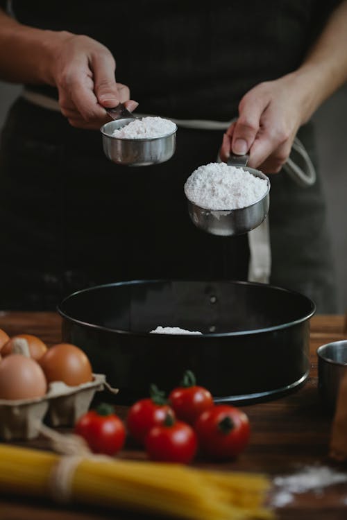 Crop unrecognizable chef in black apron standing near table and adding flour in dishware while cooking homemade pasta with eggs and spaghetti near tomatoes