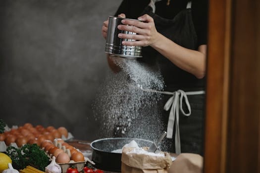 Chef sifts flour onto a counter surrounded by fresh ingredients, preparing a meal.