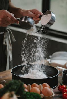 Crop anonymous chef pouring flour into baking dish while preparing ingredients for recipe with eggs and tomatoes