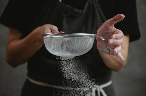 Crop anonymous cook in apron sifting flour while preparing baking dish against gray background