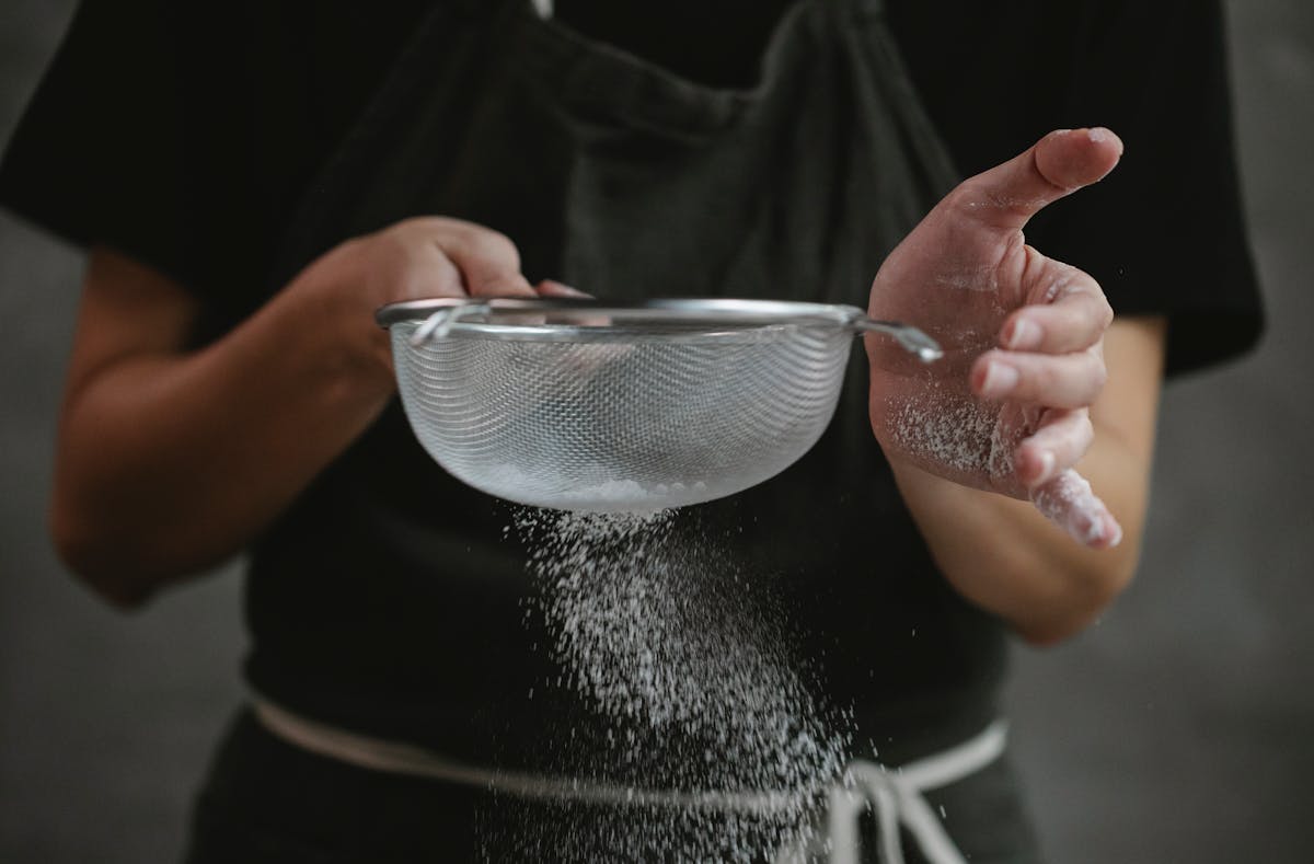 Crop anonymous cook in apron sifting flour while preparing baking dish against gray background