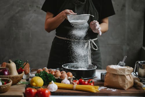 Free Crop unrecognizable cook adding flour into stainless baking dish while cooking meal with spaghetti cherry tomatoes garlic herbs and lemon Stock Photo