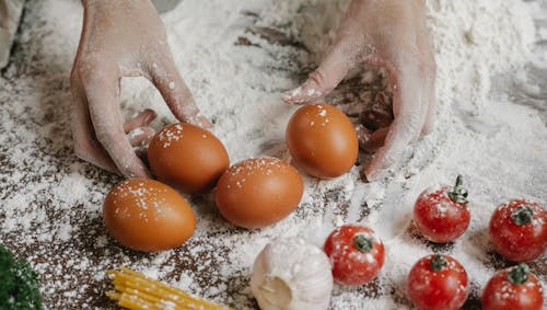 Free From above of crop anonymous female cook taking eggs while preparing meal with cherry tomatoes garlic spaghetti and flour Stock Photo