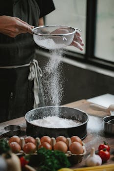 A chef sifts flour into a baking dish surrounded by fresh ingredients in a cozy kitchen.