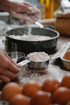 Unrecognizable cook adding flour into baking form while preparing pastry in kitchen at daylight