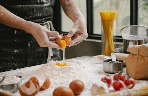 Crop anonymous person adding egg in flour while cooking pastry for recipe with cherry tomatoes and spaghetti